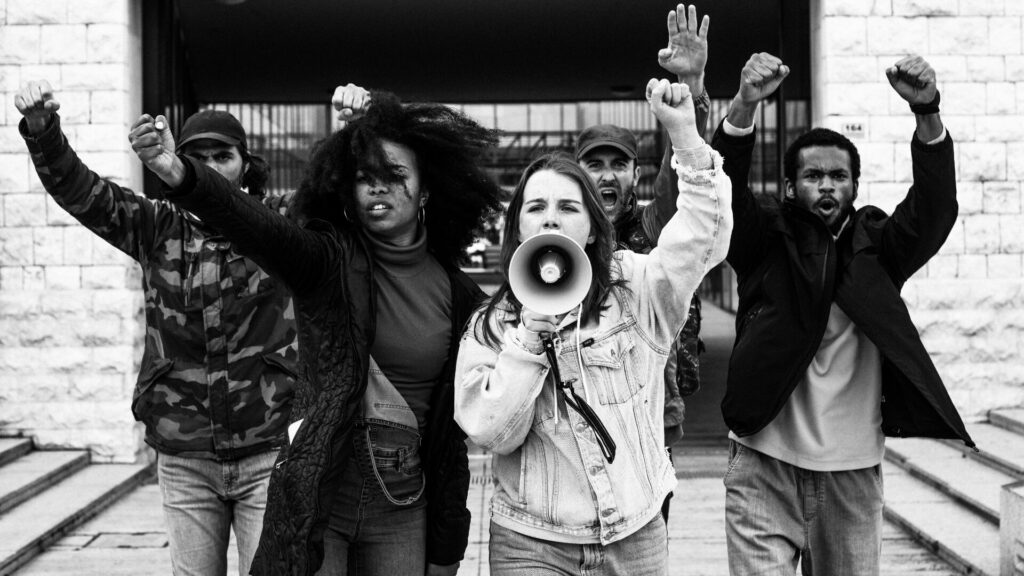 A black and white photo of a diverse group of individuals with the women in the front holding a megaphone while others have their arms raised.